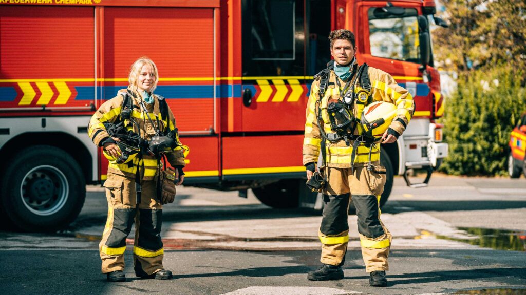 Eine Werkfeuerwehrfrau und ein Werkfeuerwehrmann stehen vor einem Feuerwehrauto, auf dem Asphaltboden sind Wasserpfützen